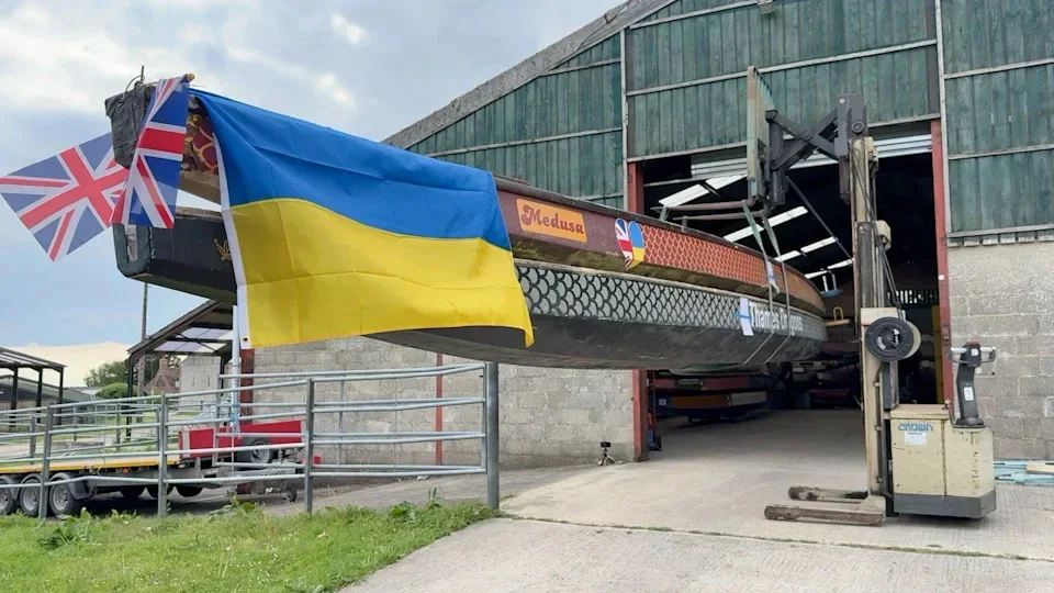 The image shows two boats lifted up by machinery. One is on top of the other. They have a Ukrainian flag and British flags on the end. The top one is red with a scale pattern on the side. It says 'Medusa' on the side. The bottom one is black with a grey scale pattern. They are half in the barn which is grey stone on the bottom and green fencing at the top.